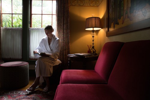 A person in a bathrobe sits on a red couch, reading a book in a warmly lit room with large windows.