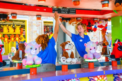 Two carnival workers celebrate at a game booth, surrounded by colorful stuffed animals and a vibrant atmosphere.