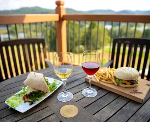 A table with a wrap, burger, fries, and two glasses of wine, set against a scenic outdoor backdrop.