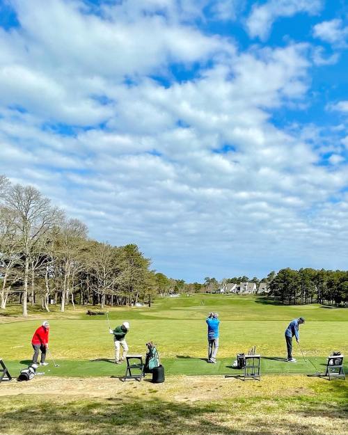 A group of golfers practicing swings on a sunny day with a blue sky and scattered clouds in the background.
