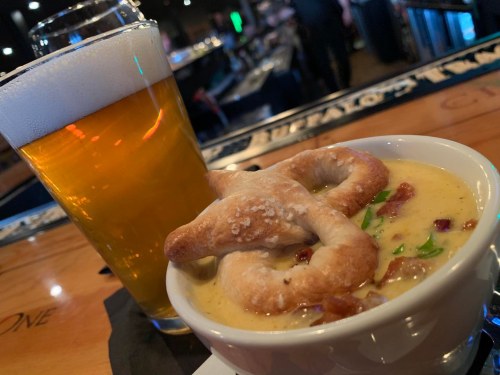 A glass of beer next to a bowl of creamy soup topped with a pretzel and garnished with green onions.
