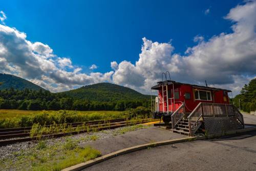 A red caboose sits beside train tracks, with green hills and a blue sky filled with clouds in the background.