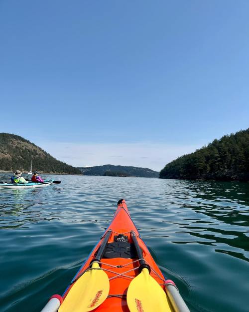 A view from a kayak on calm water, with another kayaker in the distance and lush green hills surrounding the scene.