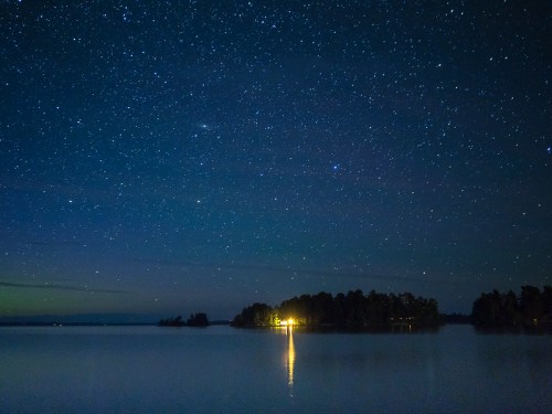 A serene night sky filled with stars above a calm lake, reflecting a distant light from an island.