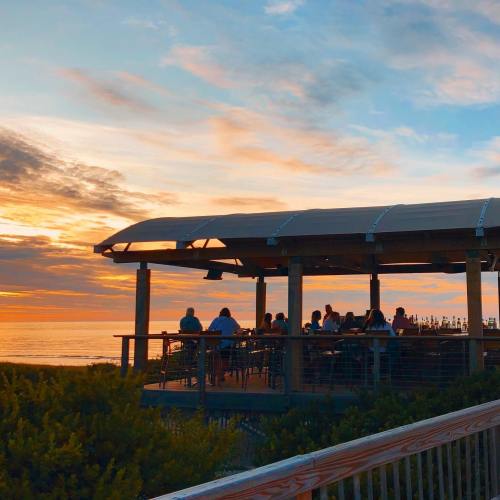 A sunset view over the ocean with people dining on a deck, surrounded by greenery.