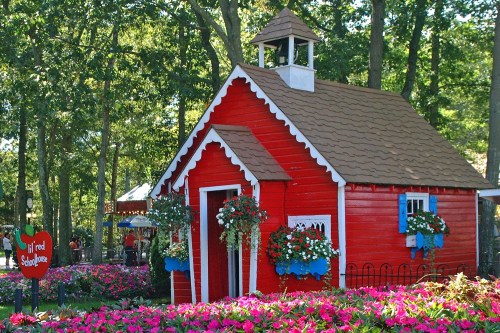 A charming red playhouse with a brown roof, surrounded by colorful flowers and greenery in a park setting.