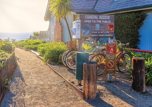 Sign for coastal access with bicycles nearby, surrounded by greenery and a pathway leading to the beach.