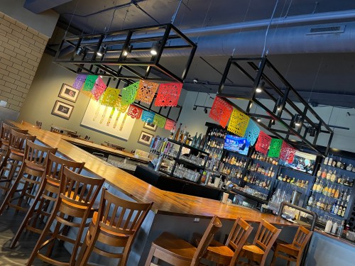 A vibrant bar area with colorful papel picado decorations hanging above, showcasing a well-stocked shelf and seating.