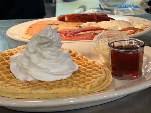 A plate with a waffle topped with whipped cream, syrup, and a side of pancakes, bacon, and eggs.