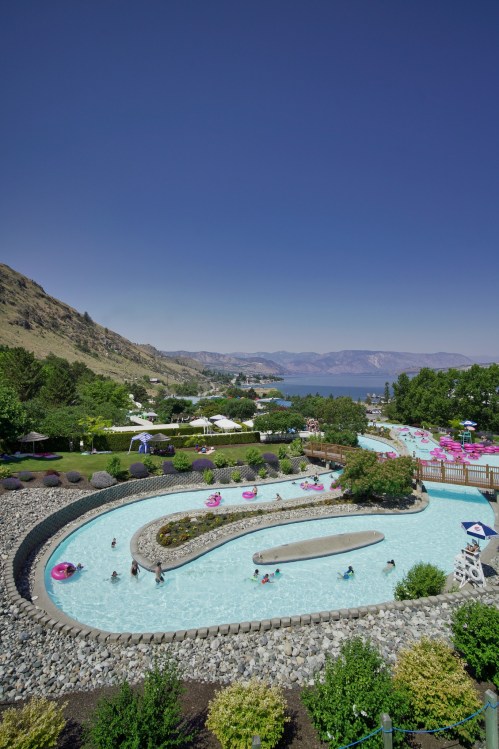 A scenic view of a lazy river pool surrounded by greenery, with mountains and a lake in the background under a clear blue sky.