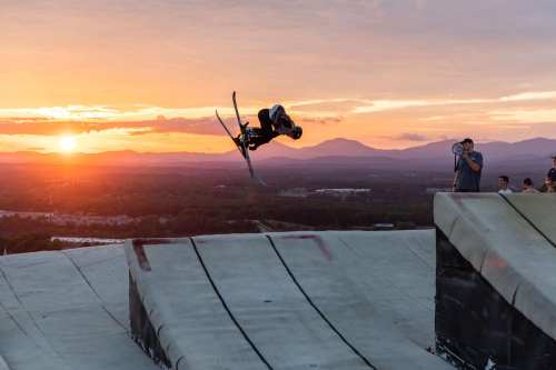 A skier performs a jump at sunset, with mountains in the background and spectators watching nearby.