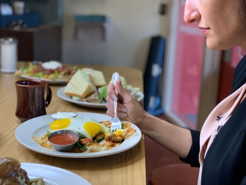 A woman holds a fork over a plate of breakfast food, with coffee and a sandwich in the background.