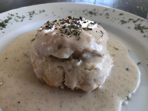 A biscuit topped with creamy gravy and herbs, served on a white plate with a light sauce around it.