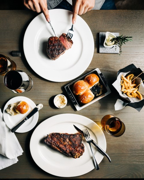 A top-down view of a dining table with steak, rolls, fries, and drinks, with hands holding utensils ready to eat.