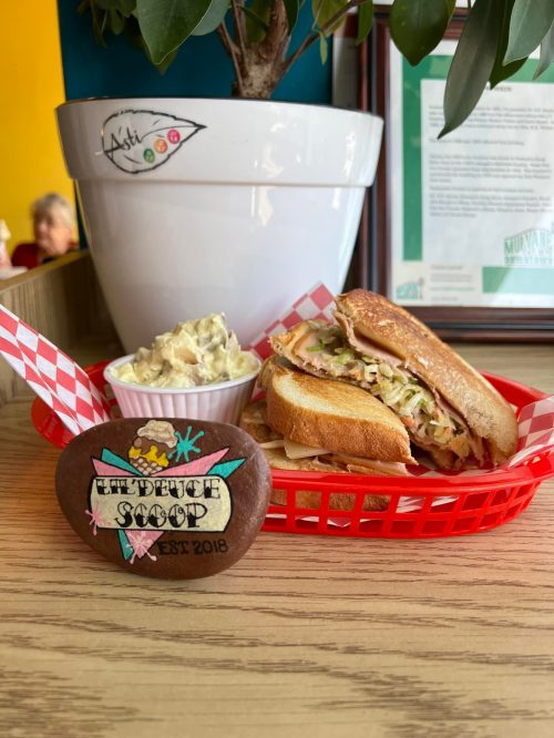A sandwich with coleslaw in a red basket, accompanied by a side of potato salad and a decorative stone.