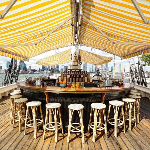 A vibrant outdoor bar with yellow-striped awnings, surrounded by stools and a view of the city skyline.