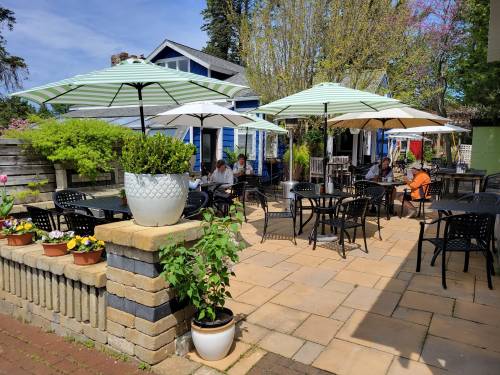 Outdoor dining area with tables under umbrellas, surrounded by greenery and flowers, in a sunny setting.