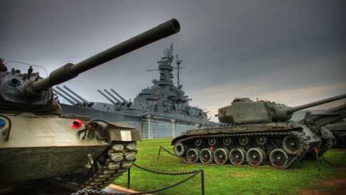 A military museum scene featuring two tanks in the foreground and a battleship in the background under a cloudy sky.