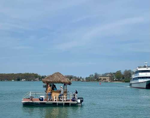 A small boat with a thatched roof floats on calm water, with a larger vessel nearby and a scenic shoreline in the background.