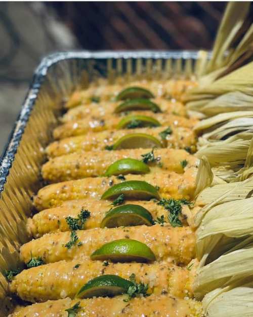 A tray of corn on the cob topped with sauce, lime slices, and herbs, with husks arranged on the side.