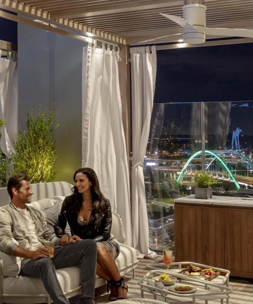 A couple enjoys drinks and snacks on a stylish balcony with city lights and a bridge in the background at night.