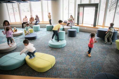 Children play on colorful seating in a bright, spacious room with large windows and adults in the background.