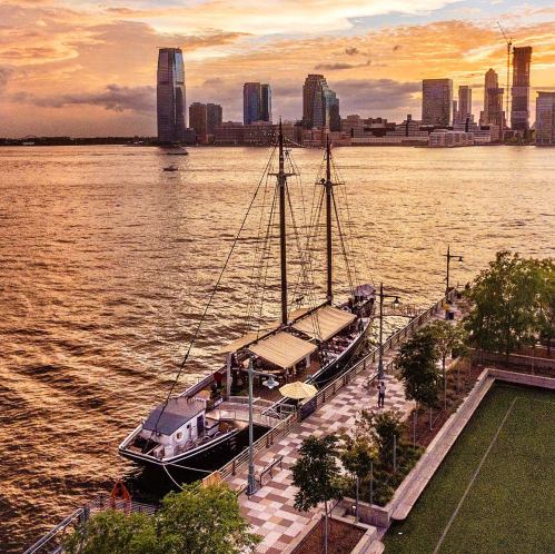 A sailboat docked near a waterfront park, with a city skyline and colorful sunset in the background.