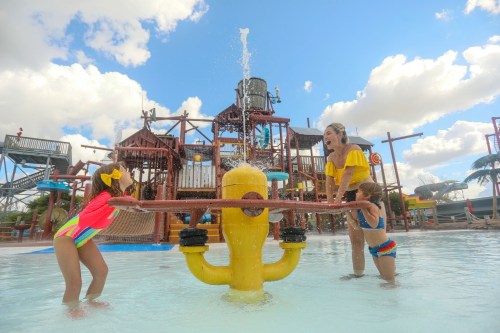 Two children play with a water feature at a splash park, while an adult smiles nearby under a bright blue sky.