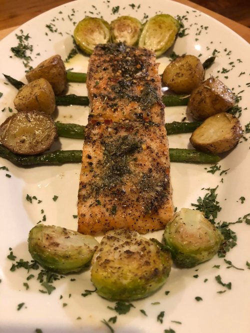 A plated dish featuring grilled salmon, roasted Brussels sprouts, and potatoes, garnished with herbs.