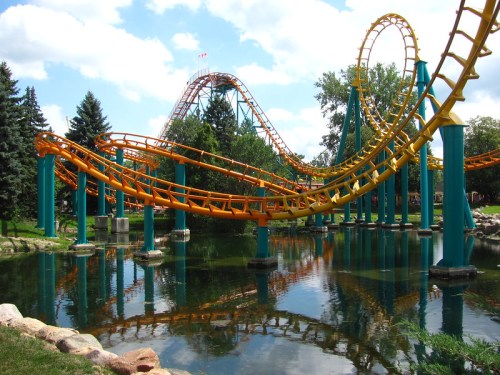 A colorful roller coaster with orange tracks twists above a calm pond, surrounded by trees and blue skies.