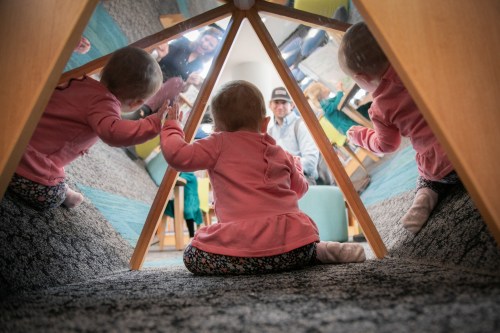 A baby sits inside a play tent, looking at reflections of themselves and adults in the mirrored surfaces.