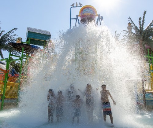 A group of children and adults stand in a splash zone at a water park, surrounded by a large burst of water.