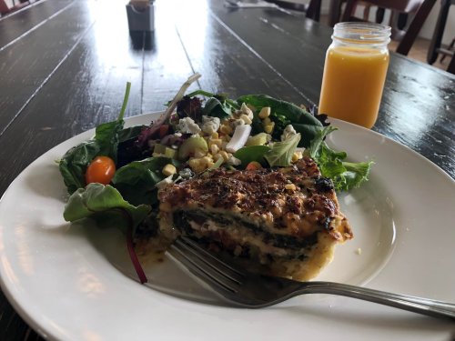 A plate with a slice of baked dish, a fresh salad, and a small jar of orange juice on a wooden table.