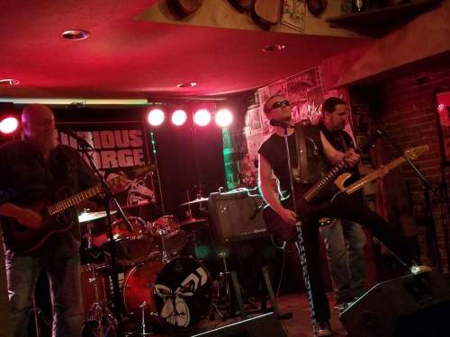 A rock band performs on stage with colorful lights, featuring a guitarist, bassist, and a lead singer in sunglasses.