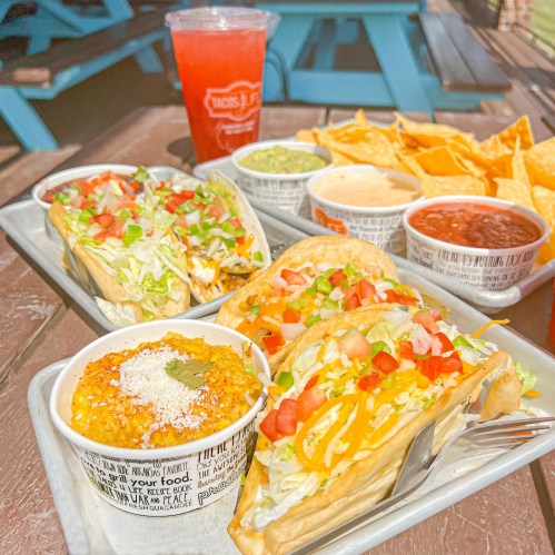 A colorful meal featuring tacos, sides of guacamole, queso, salsa, and tortilla chips, with a drink on the side.