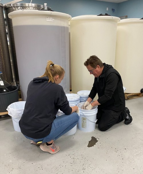 Two people, one kneeling and one squatting, are working with buckets in a room with large storage tanks.