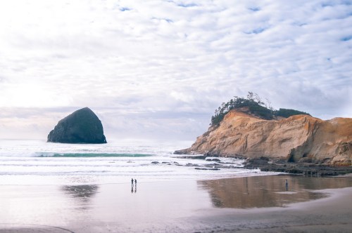 Two people walk along a sandy beach with a large rock formation and cliffs in the background under a cloudy sky.