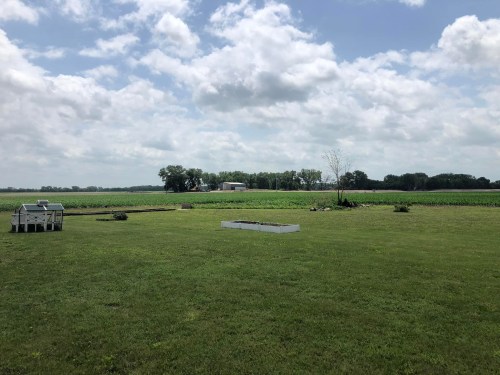A grassy field with a few trees, a small structure, and a distant farm under a partly cloudy sky.