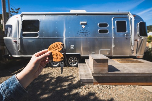 A hand holds an orange keychain in front of a shiny silver Airstream trailer and a wooden deck.