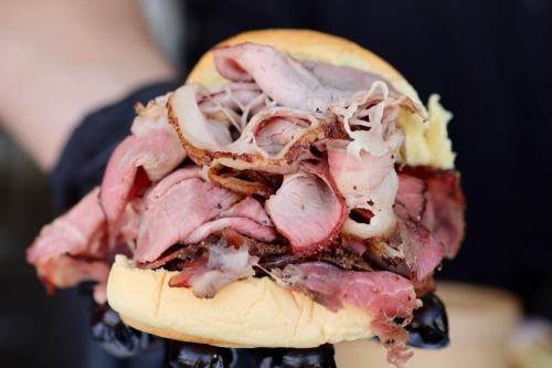 A close-up of a large sandwich filled with layers of various meats, held in a gloved hand.