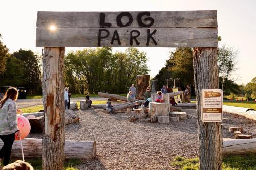 A wooden sign reads "Log Park" at a playground with children and adults playing on logs and enjoying the outdoors.