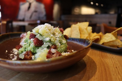 A bowl of guacamole topped with cheese and bacon, served with a side of tortilla chips on a wooden table.