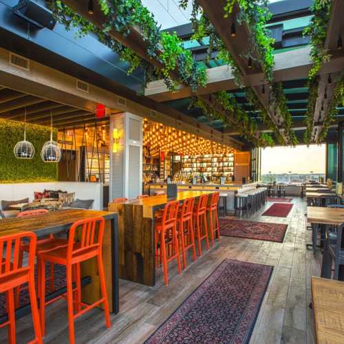 A modern restaurant interior with orange chairs, wooden tables, and greenery, featuring a bar and large windows.