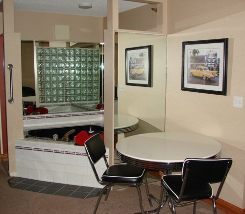A small room featuring a round table with two chairs, a glass block wall, and framed car photos.