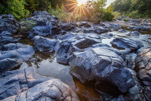 Sunlight beams over rocky terrain with pools of water, surrounded by lush greenery and a serene landscape.