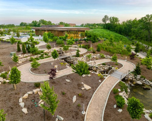 A landscaped garden with winding paths, trees, and a modern building in the background under a clear sky.