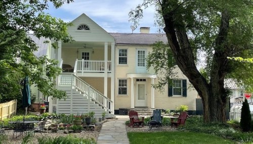A charming two-story house with a porch, surrounded by greenery and a stone pathway leading to red chairs in the yard.