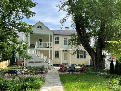 A charming two-story house with a porch, surrounded by greenery and a stone pathway leading to a garden seating area.