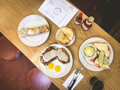A wooden table with plates of breakfast: eggs, toast, a cinnamon roll, and condiments.