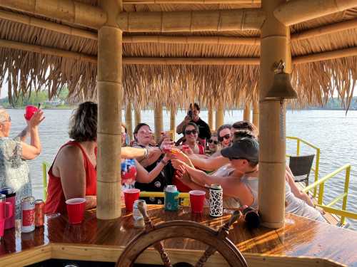 A group of friends enjoying drinks under a thatched roof by the water, smiling and celebrating together.